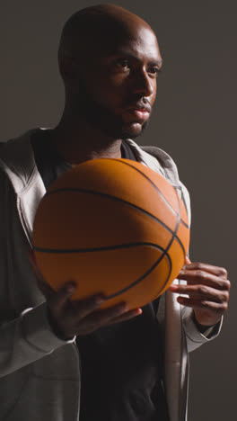 Vertical-Video-Studio-Vertical-Video-Portrait-Shot-Of-Male-Basketball-Player-Wearing-Tracksuit-Throwing-Ball-From-One-Hand-To-The-Other-Against-Dark-Background